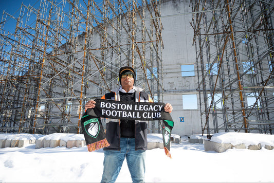 A man stands in front of a partially constructed building, holding a black scarf that reads 'BOSTON LEGACY FOOTBALL CLUB.' He is wearing a winter jacket and a hat, with snow covering the ground around him. The background features scaffolding and a clear b
