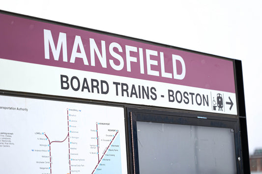 A close-up of a train station sign reading 'MANSFIELD' in large white letters. Below, the sign states 'BOARD TRAINS - BOSTON' with an arrow pointing right, accompanied by a train icon. The lower section features a map of the train routes in the area. The 
