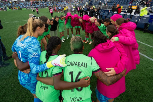 Boston Legacy FC players and staff huddle together on the pitch before or after a match, arms around each other in a circle, wearing green kits and pink team jackets.