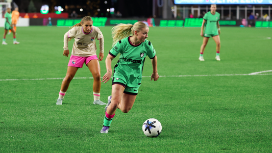 Boston Legacy FC player, Chloe Ricketts, drives forward with the ball during a night match, pursued by an opposing player in a cream and pink kit.
