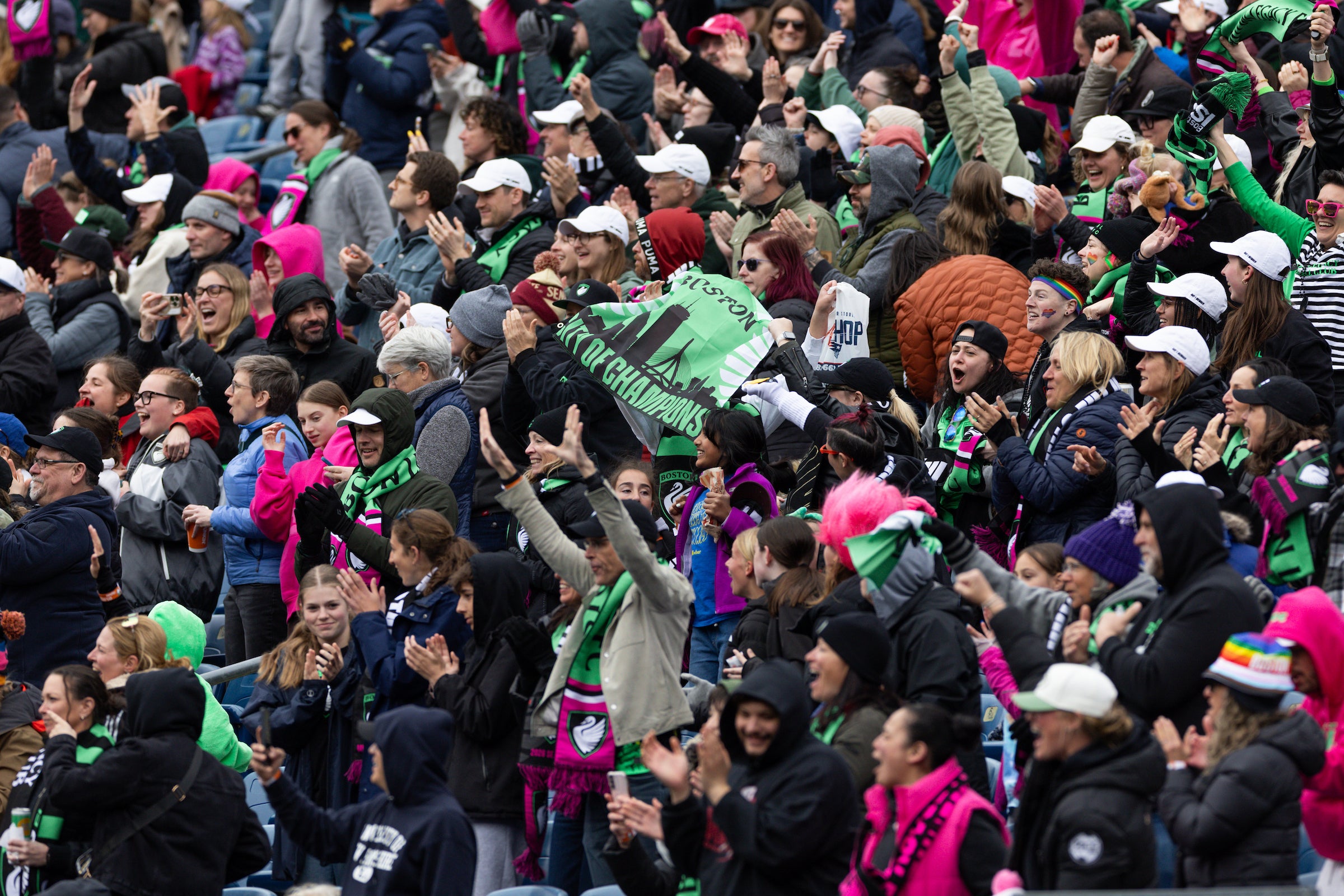 A packed supporters’ section erupts in celebration at a soccer match, with fans wearing team scarves, winter jackets, and bright pink and green accessories. Many people are cheering with raised arms and clapping, while one fan waves a green banner in the center of the crowd. The atmosphere feels energetic and communal, with the stands filled by a diverse mix of supporters enjoying the game together.