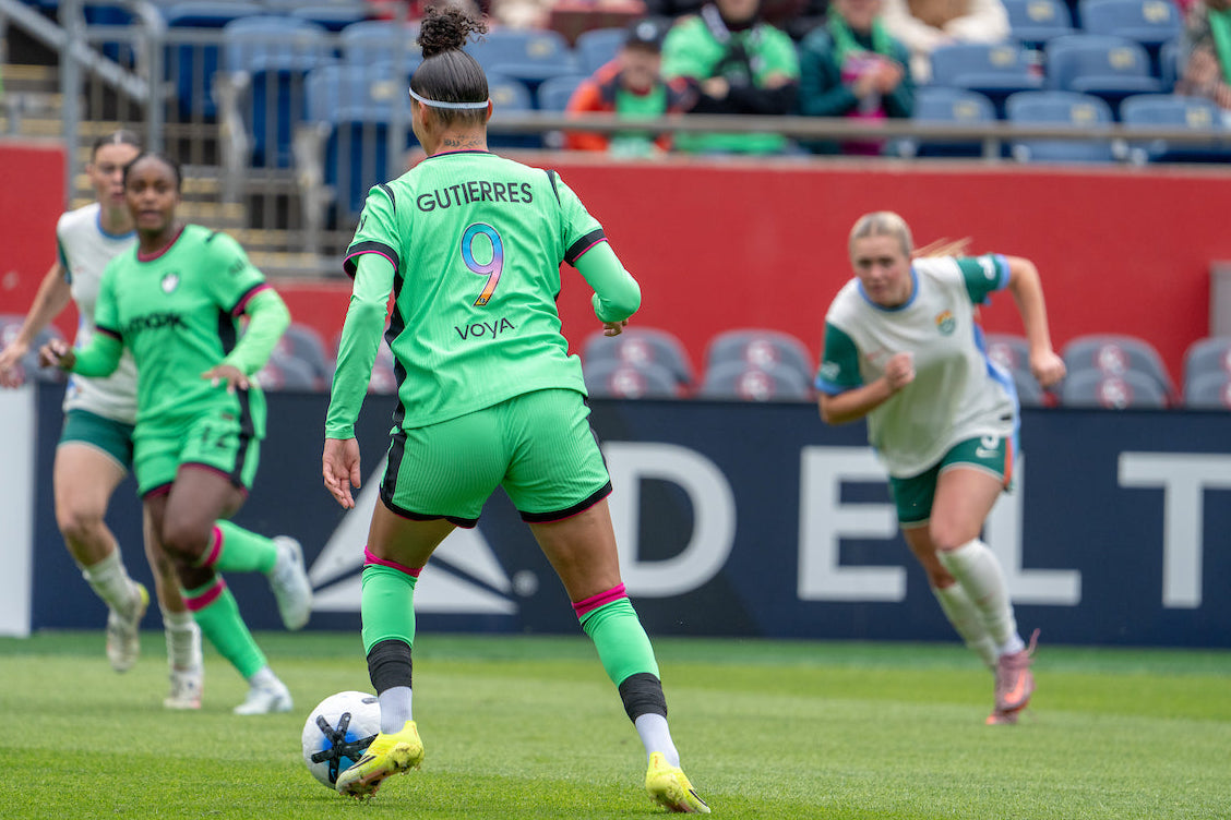 A soccer player wearing a bright green long-sleeve jersey with 'GUTIERRES' and the number 9 on the back, standing on a green field and controlling a soccer ball. Other players in green and white are visible in the background, along with stadium seating and a blue advertising board.