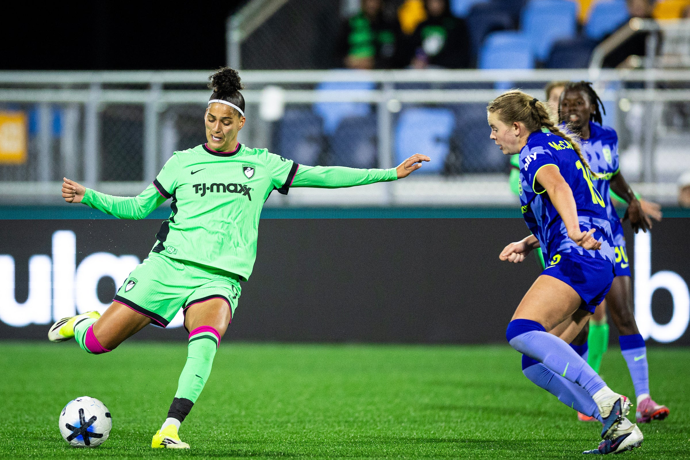 A soccer player in a bright green uniform, branded with TJ Maxx, skillfully kicking the ball while being challenged by a player in a blue uniform. The action takes place on a well-lit field, with spectators visible in the background, capturing the intensity of the match.