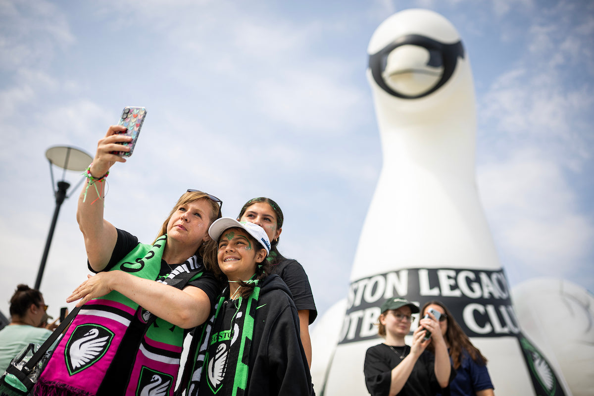 Boston Legacy FC fans take a selfie in front of a large inflatable swan mascot branded with the Boston Legacy Football Club logo at an outdoor pregame event.