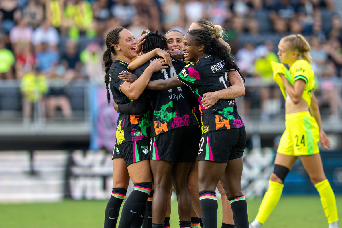 A group of female soccer players in black jerseys with vibrant neon green, pink, and orange graphics celebrate a goal on a green field, with a teammate in a bright neon yellow uniform standing nearby.