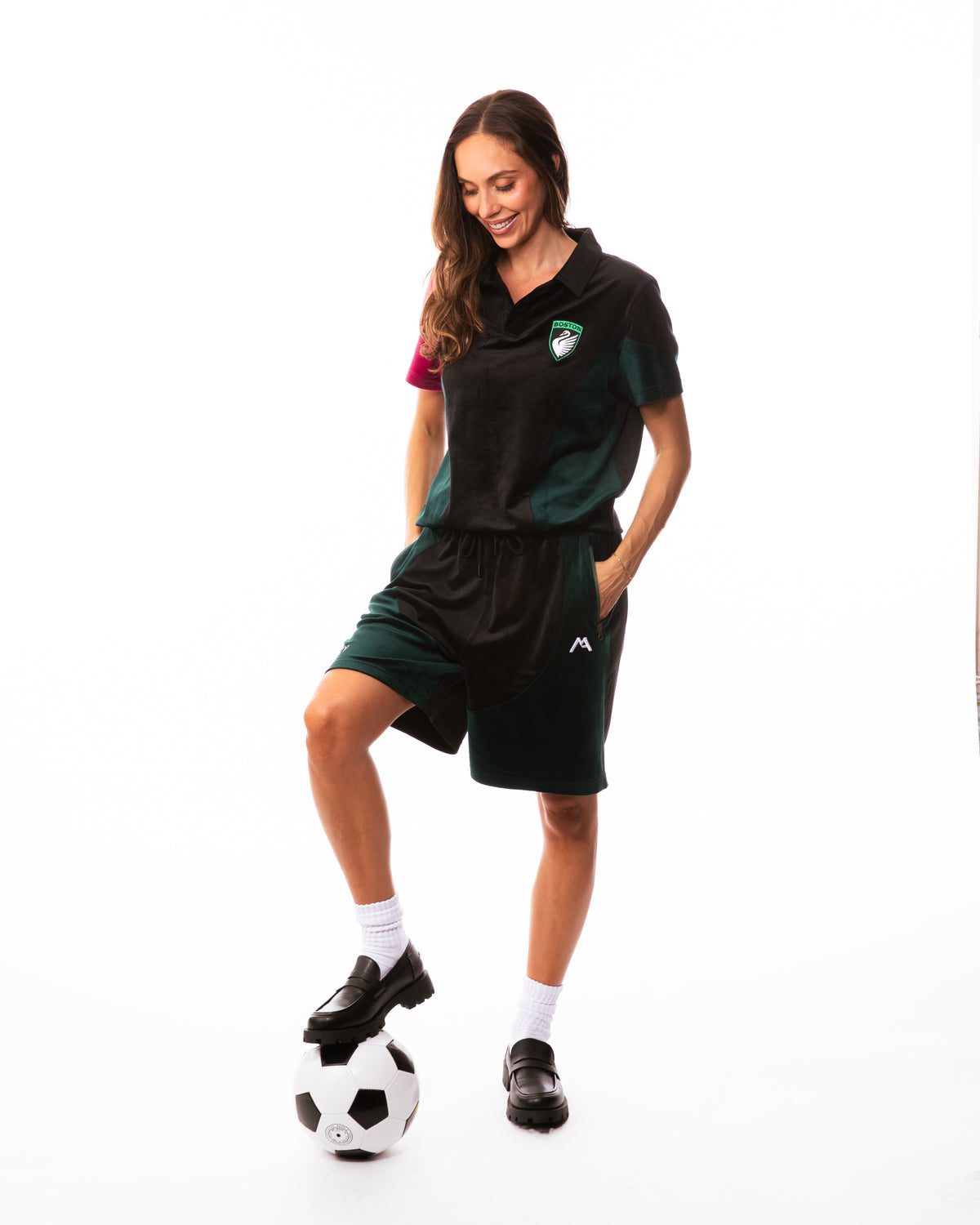 A woman modeling a dark green and black athletic soccer polo shirt and matching shorts, balancing a black and white soccer ball on her foot against a white studio background.